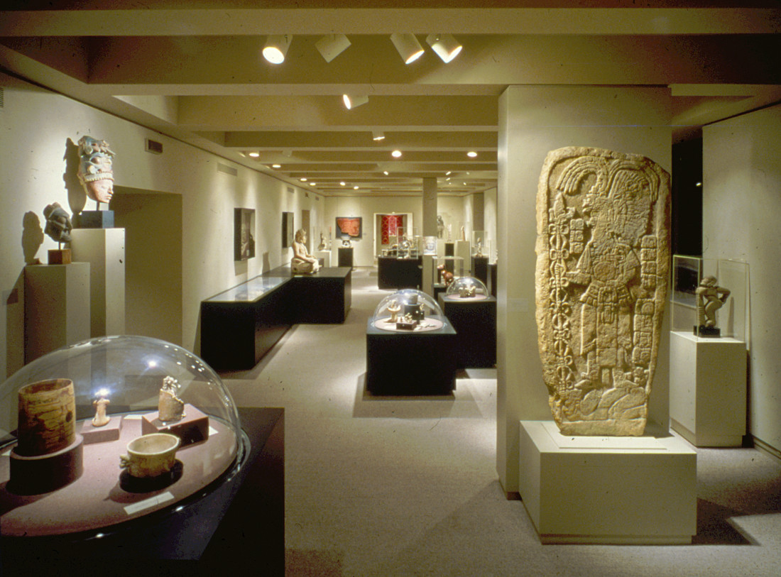 A color installation photograph of a gallery. There are multiple pedestals and cases with domed glass covering ancient American artifacts.