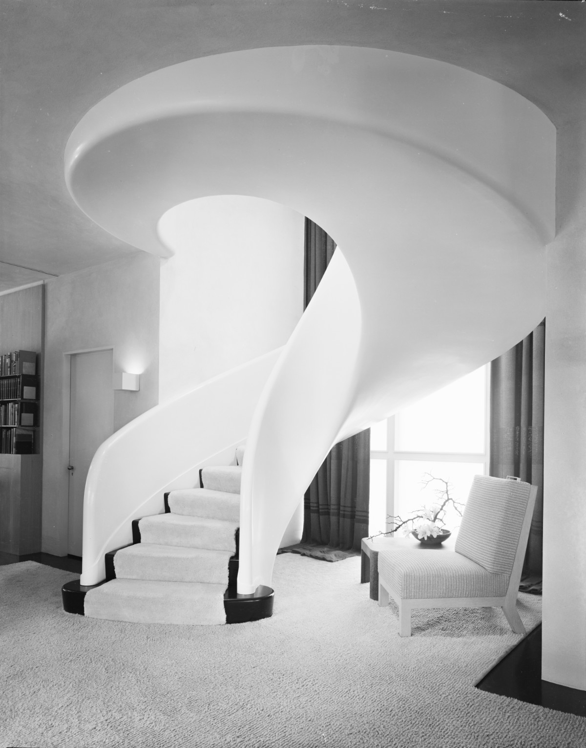 A black-and-white photograph of a house interior with a white carpeted spiral staircase. There is a chair next to the window beneath the stairs.