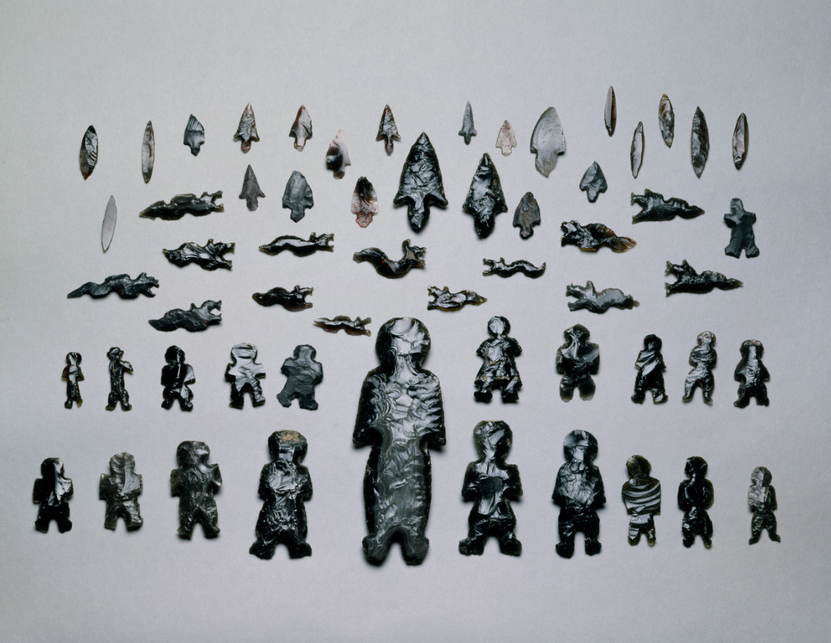A color photograph of fifty-eight black obsidian objects arranged on a table. Twenty-one are humanoid shaped of varying sizes. Fourteen resemble snakes with open mouths, fifteen are standard arrowheads, and seven are shards.