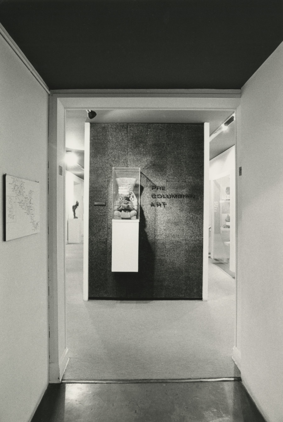 A black-and-white photograph of a hallway leading into a gallery space, where there is a dark free-standing wall with the title, “Pre Columbian Art” on it and  an attached case that holds a ceramic seated figure.