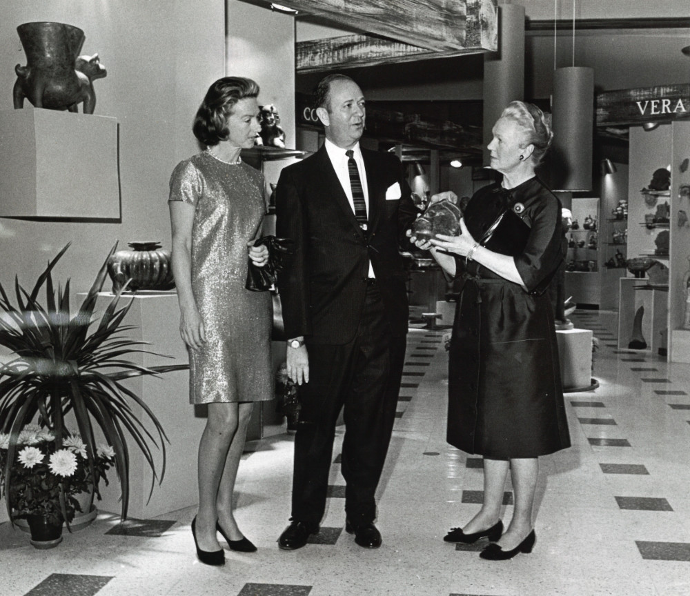 A black-and-white photograph of two well-dressed women and a man standing in department store. The woman on the right holds a stone mask. Behind them, shelves and pedestals display ancient American objects.