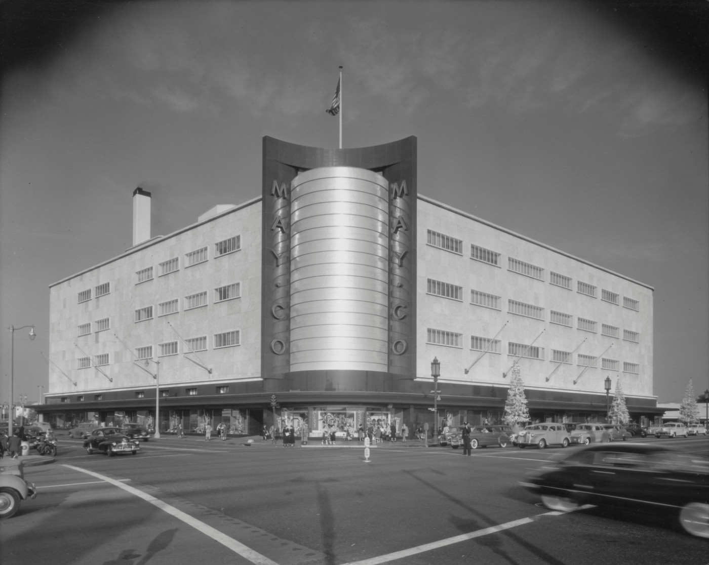 A black-and-white photograph of a four-storied May Co. department store on the corner of a busy intersection.