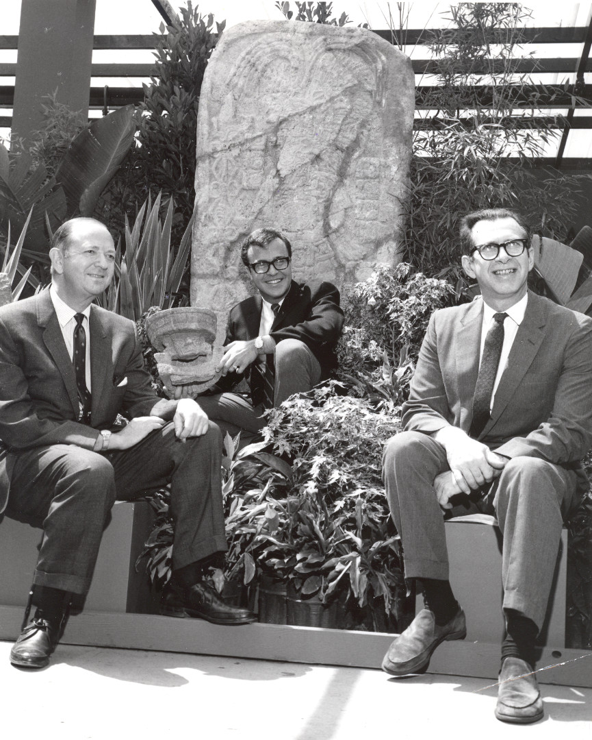 A black-and-white photograph of three men in suits seated in front of a monumental stone carving surrounded by plants. The man in the middle holds a stone object in his hand.