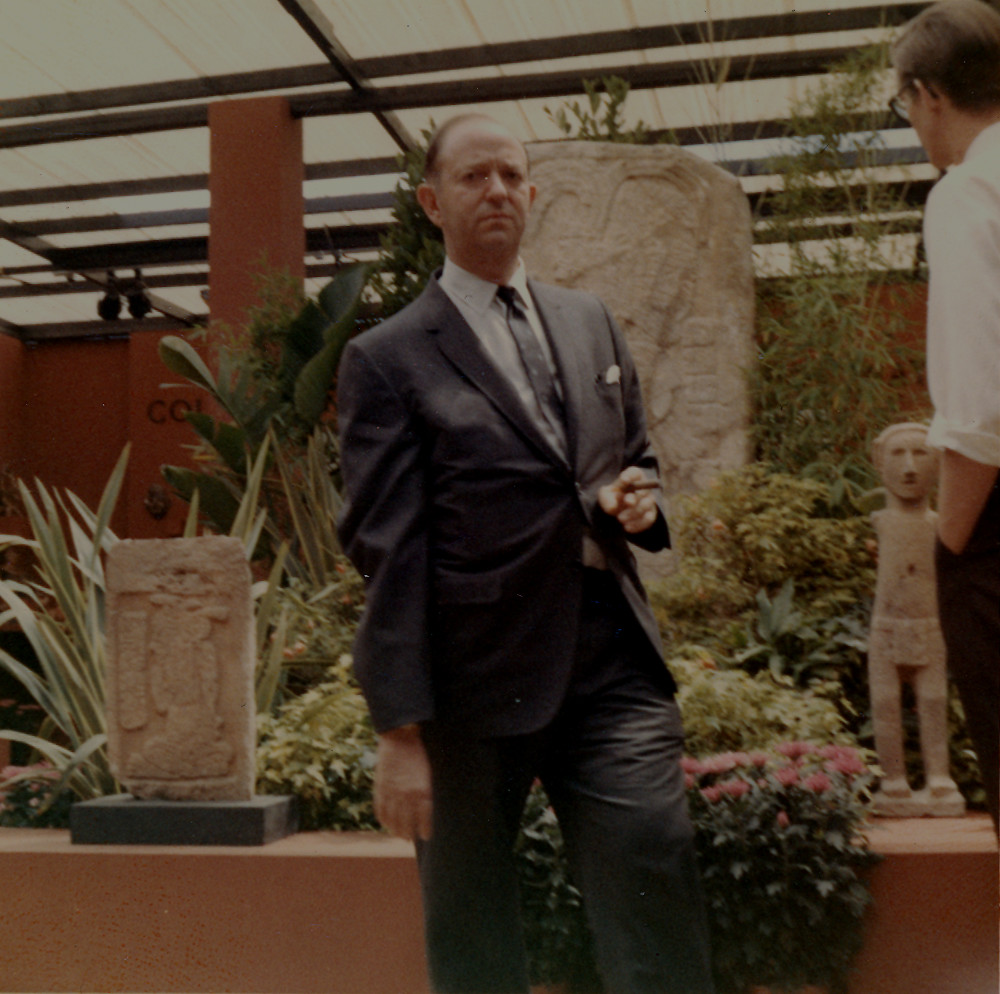 A color photograph of a man in a suit standing outside in front of a pedestal with two ancient American sculptures and a monumental stone carving among foliage.