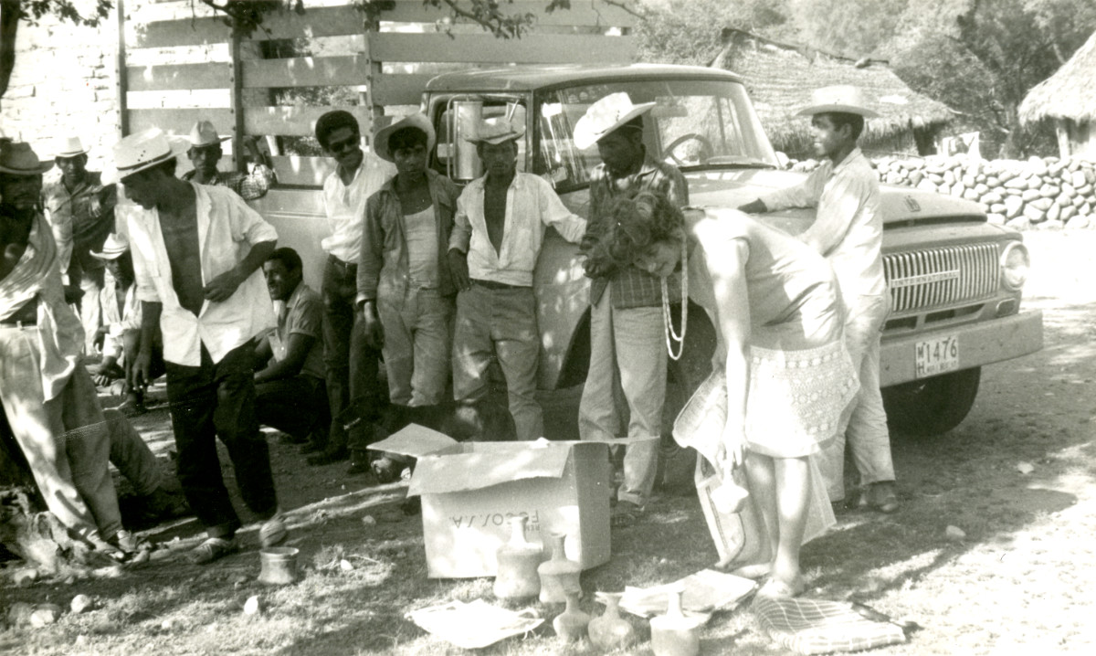 A black-and-white photograph of ten men in work attire standing next to a truck on an unpaved road. A woman in a dress leans over a group of ceramic objects, holding a newspaper page in one hand and a ceramic in the other.